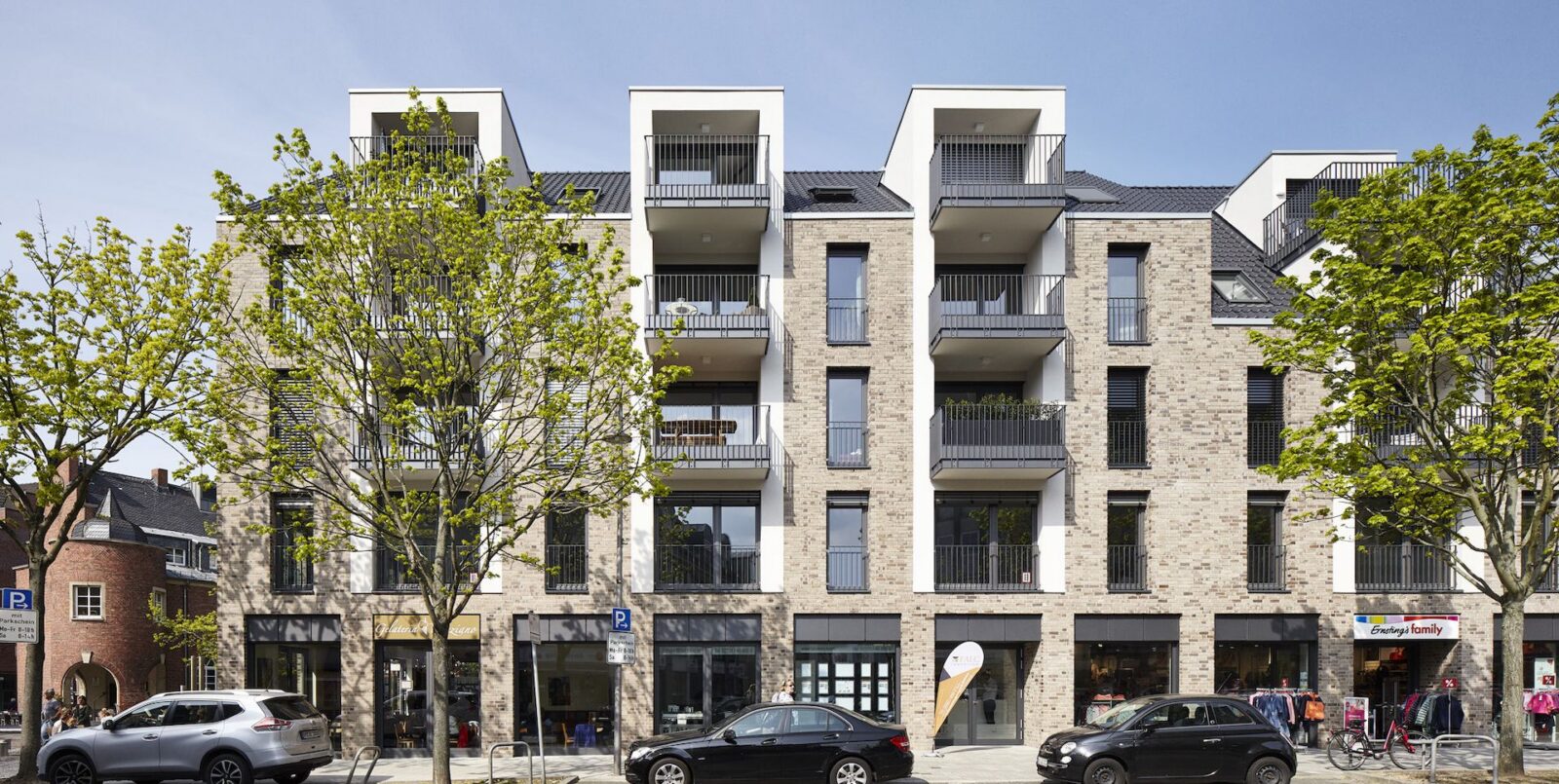 Ein modernes Wohnhaus mit großen Balkonen, hohen Fenstern und einer Backsteinfassade. Bäume und geparkte Autos säumen die Straße davor; Geschäfte und ein Lebensmittelgeschäft befinden sich im Erdgeschoss.