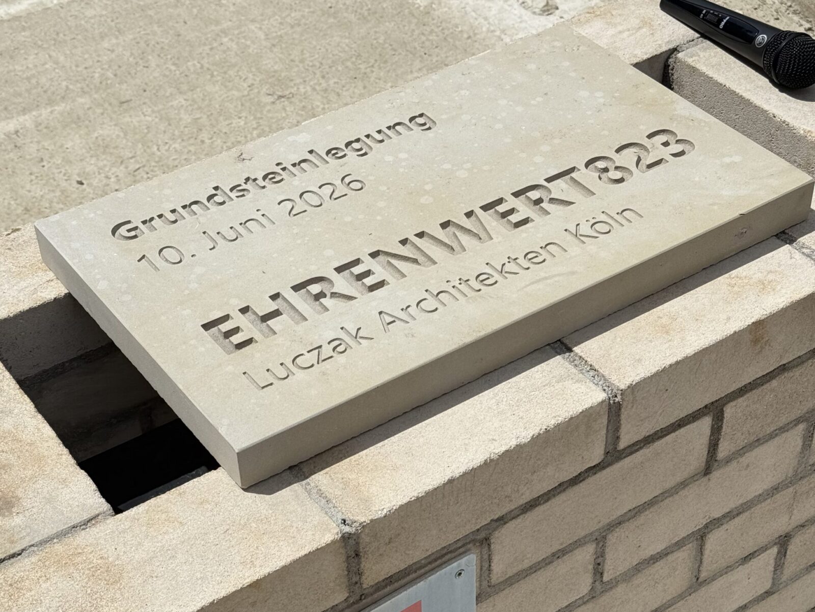 Auf einem Backsteinsockel ruht ein Grundstein, auf dem „EHRENWERT823“, das Datum „10. Juni 2026“ und „Luczak Architekten Köln“ eingraviert sind. Auf einer darunter liegenden Gedenktafel steht „Friedrich Wassermann“. In der Nähe liegt ein Mikrofon auf dem Boden.
