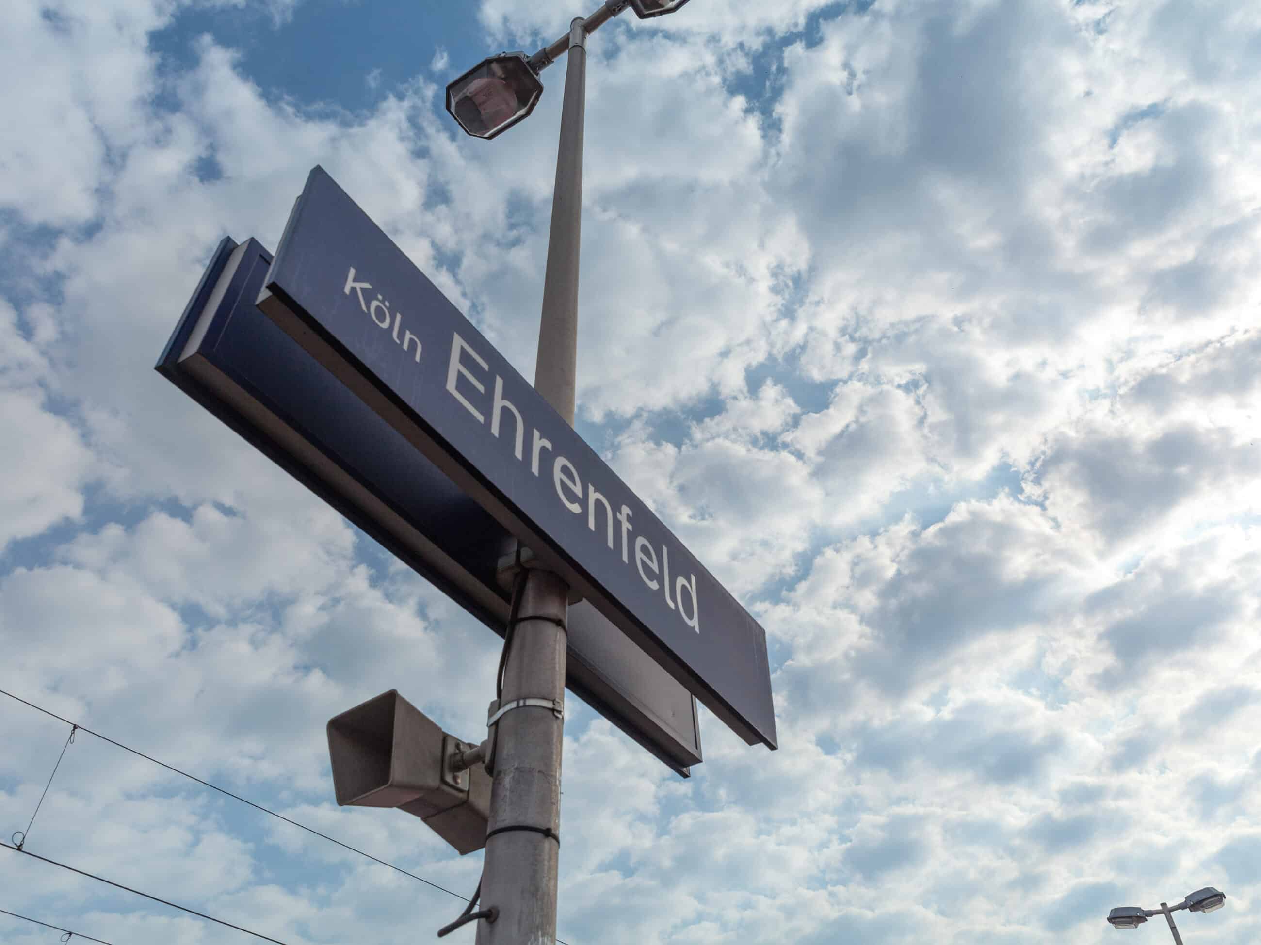 Ein Straßenschild für Köln Ehrenfeld ist unter einem teilweise bewölkten Himmel an einem Mast befestigt, im Hintergrund sind Stromleitungen und Straßenlampen zu sehen.