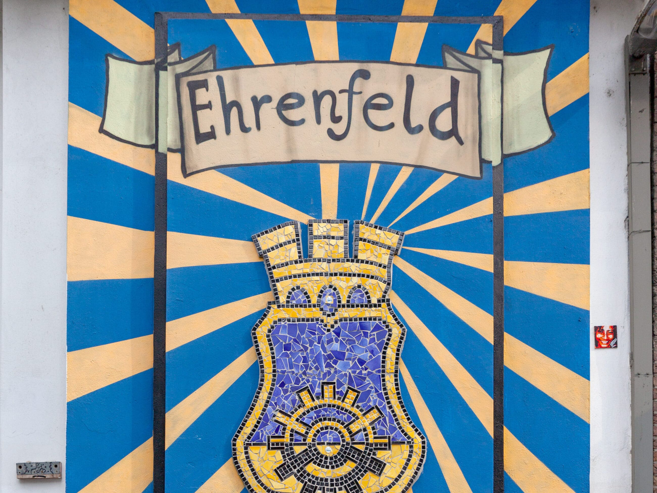 Ein Wandgemälde mit einem Banner mit dem Wort "Ehrenfeld" über einem gelben und blauen Mosaikwappen vor einem blauen Hintergrund mit gelben Strahlen, die nach außen strahlen.