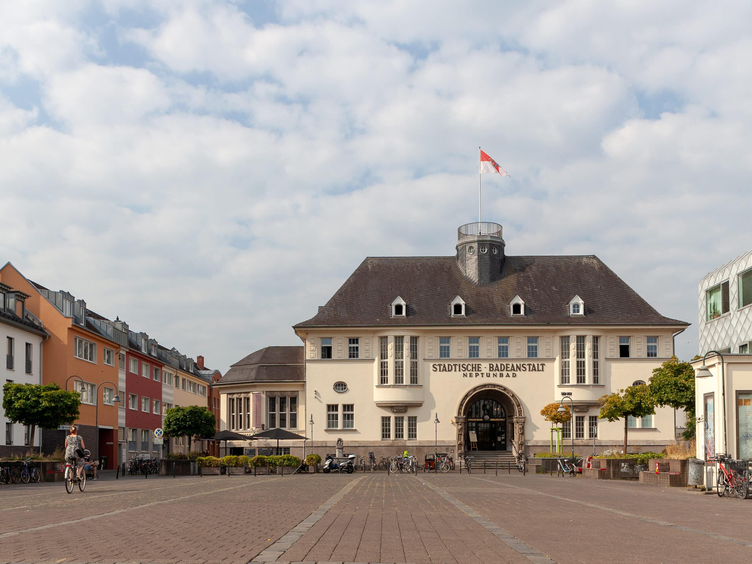 Ein historisches Gebäude mit einem zentralen Turm und einer Schweizer Fahne auf der Spitze, das die Bezeichnung Städtische Badeanstalt trägt, steht am Ende eines breiten Kopfsteinpflasterplatzes. Auf beiden Seiten des Platzes stehen bunte Gebäude, auf denen Menschen radeln und spazieren gehen.