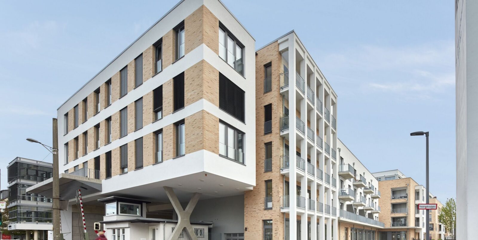 Ein modernes mehrstöckiges Wohnhaus mit hellem Backstein und weißen Akzenten, mit großen Fenstern und Balkonen, unter einem klaren blauen Himmel. Die untere Etage hat einen einzigartigen Überhang, der von schrägen Säulen getragen wird.