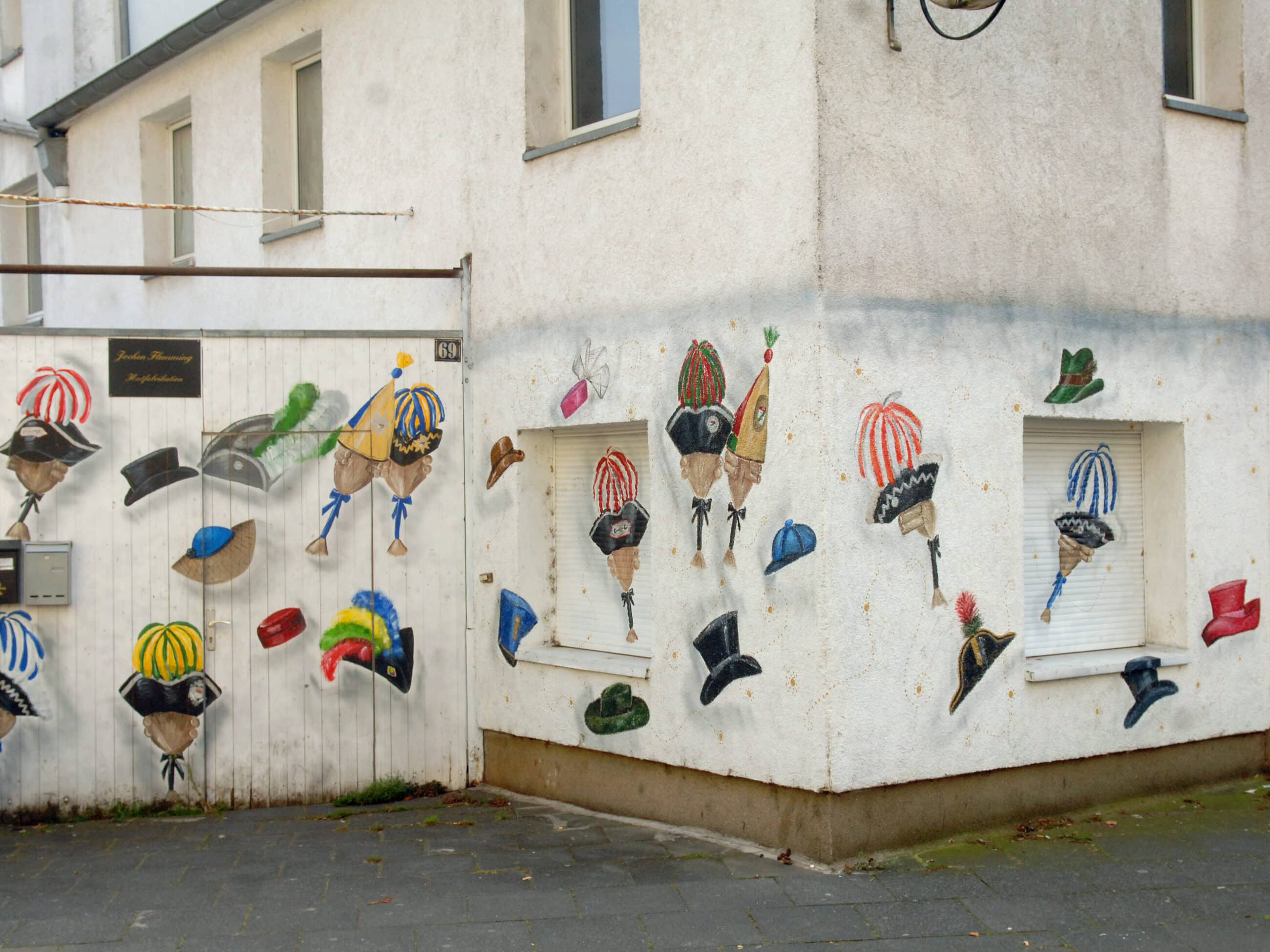 Weiße Gebäudeecken mit bunten, skurrilen Hüten und Masken an den Wänden und Fenstern; einige Hüte sind gestreift, gefedert oder mit Accessoires verziert. Die Straße und das Gebäude wirken sauber, aber verwittert.