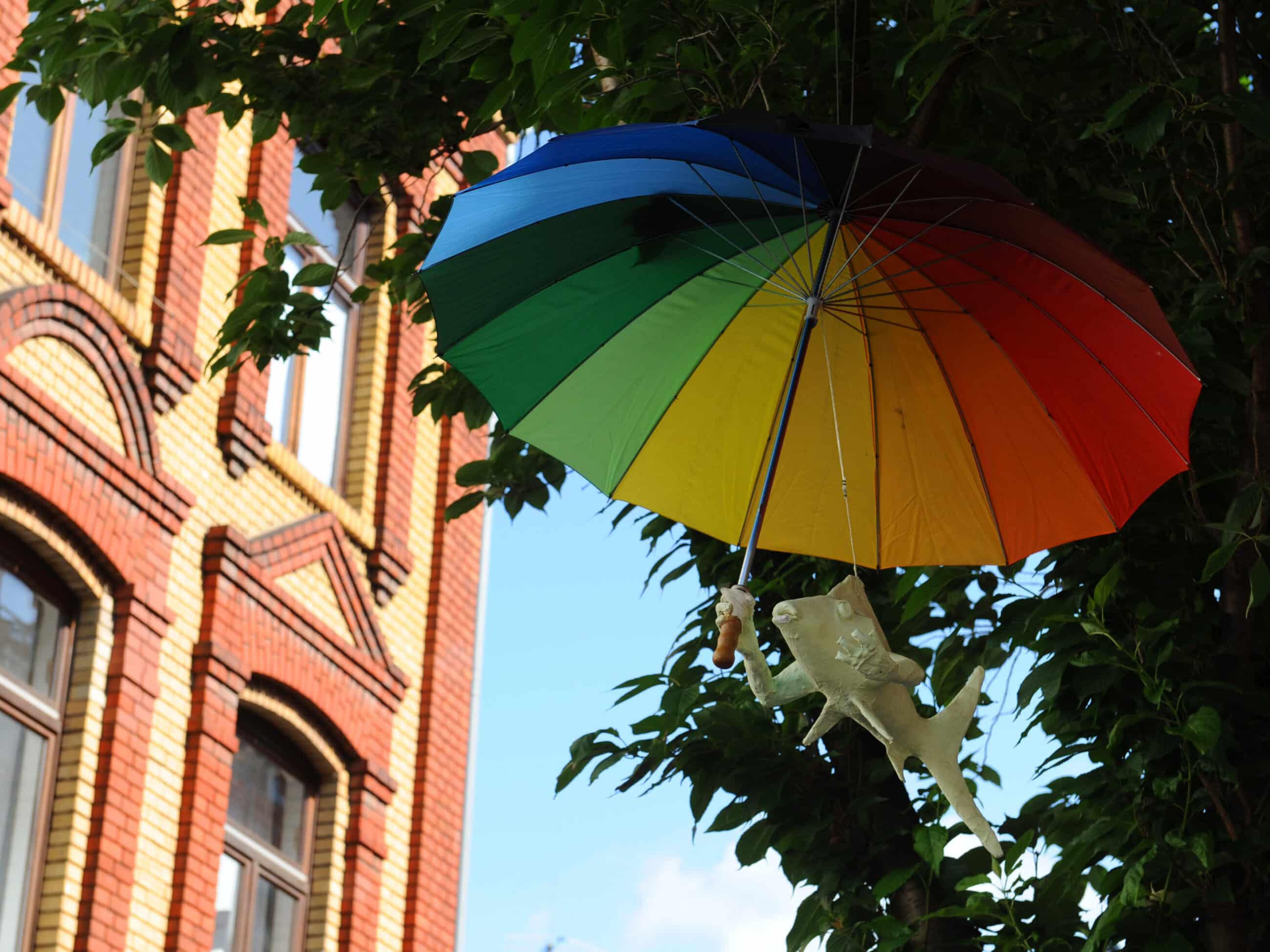 Ein regenbogenfarbener Regenschirm hängt an einem Baum neben einem Backsteingebäude. Unter dem Schirm schwebt eine kleine Dinosaurierfigur in der Luft.