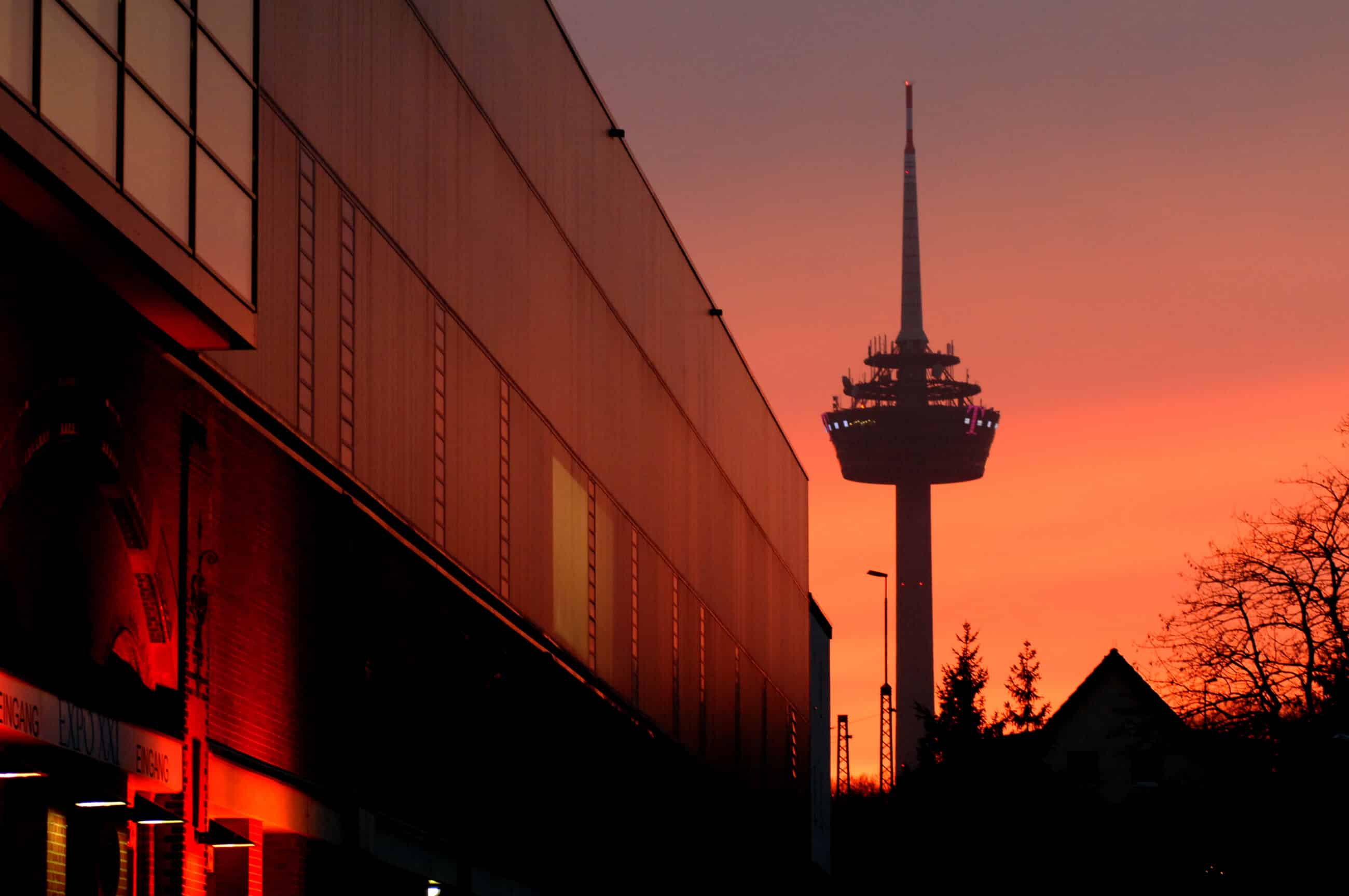 Ein hoher Fernmeldeturm steht vor einem leuchtend orangefarbenen Sonnenuntergangshimmel, mit Silhouetten von Gebäuden und Bäumen im Vordergrund und einem modernen Gebäude, in dem sich das Licht des Sonnenuntergangs spiegelt, auf der linken Seite.